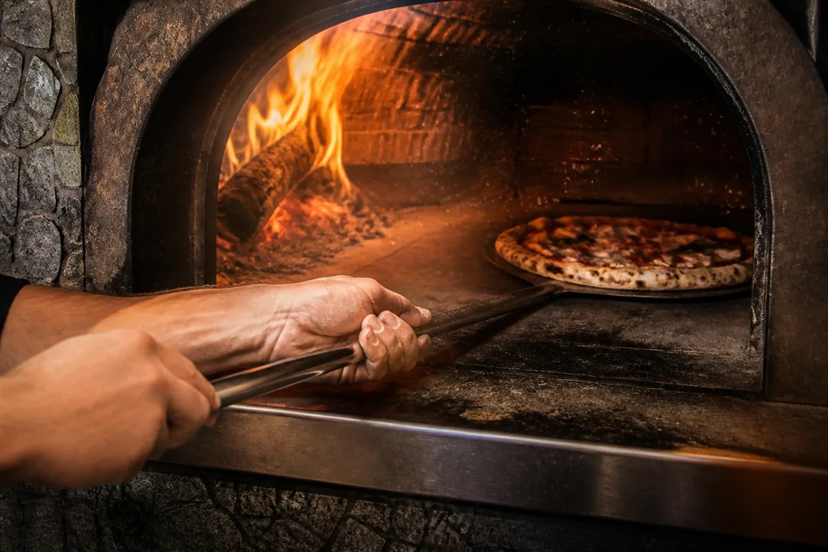 Bouche d’un four à pizza en dôme, braises rougeoyantes sur le côté, pizza posée sur la sole en pierre claire